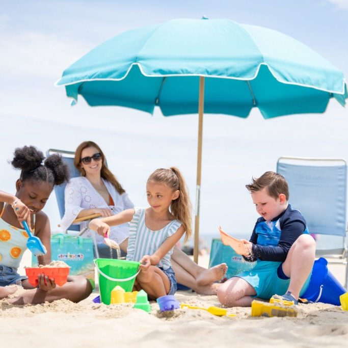 A family enjoys fun in the sun at the beach located on Jenkinson's Boardwalk. A woman sits in a beach chair under an umbrella and next to a Jenkinson's cooler bag. Three kids sit and play in the sand with plastic toys and buckets